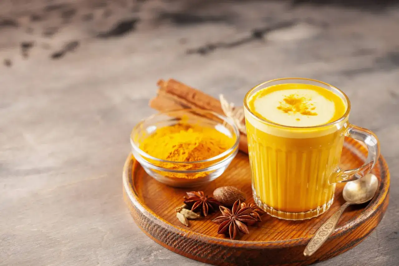 Goldene Milch: Ein Glas mit leuchtend gelbem Getr&auml;nk, daneben eine Schale Kurkumapulver, Zimtstangen und Gew&uuml;rze. Perfekt f&uuml;r ein w&auml;rmendes goldene milch rezept.