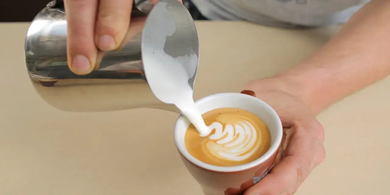 Milchschaum flie&szlig;t aus einem K&auml;nnchen in eine Tasse mit Kaffee. So einfach ist es, einen Cappuccino selber zu machen und ein sch&ouml;nes Muster zu kreieren.