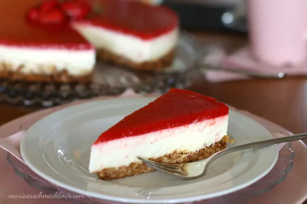 Ein St&uuml;ck New York Cheesecake ohne Backen mit roter Fruchtschicht auf einem Teller, daneben ein ganzer Kuchen.