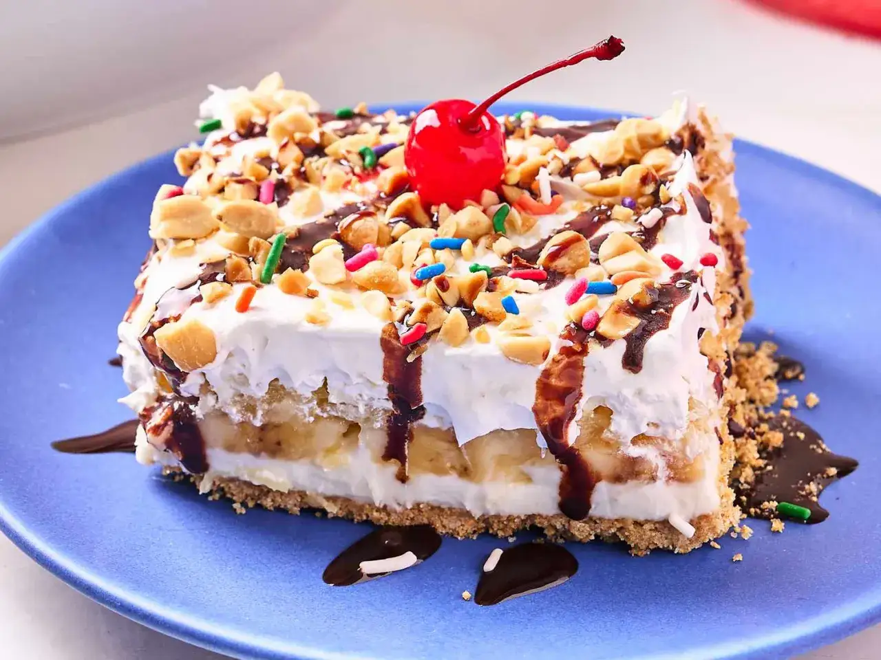 Ein St&uuml;ck Bananensplit Torte ohne Backen, dekoriert mit N&uuml;ssen, Schokosauce und einer Kirsche.