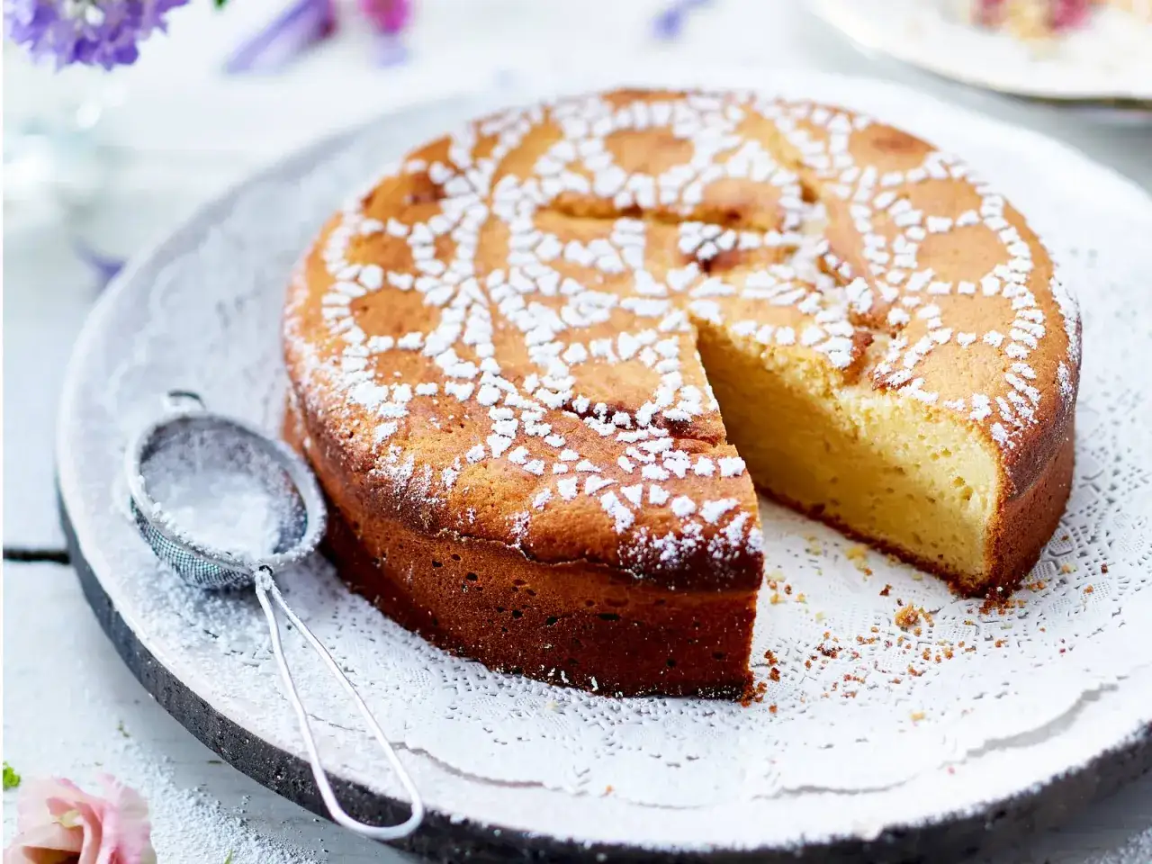 Ein goldbrauner, saftiger Omas Joghurt Kuchen, best&auml;ubt mit Puderzucker, daneben ein Sieb.