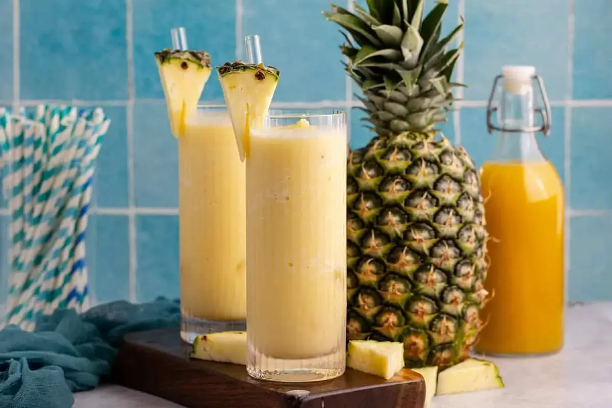 Zwei erfrischende Gl&auml;ser alkoholfreier Pi&ntilde;a Colada, garniert mit Ananasst&uuml;cken, neben einer frischen Ananas und einer Flasche Saft.