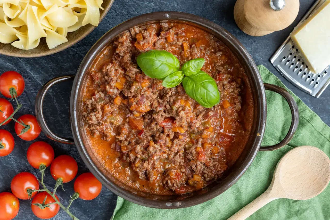 Herzhaftes Bolognese-Rezept original in einem Topf, garniert mit Basilikum. Daneben frische Tomaten, Bandnudeln und Parmesan.