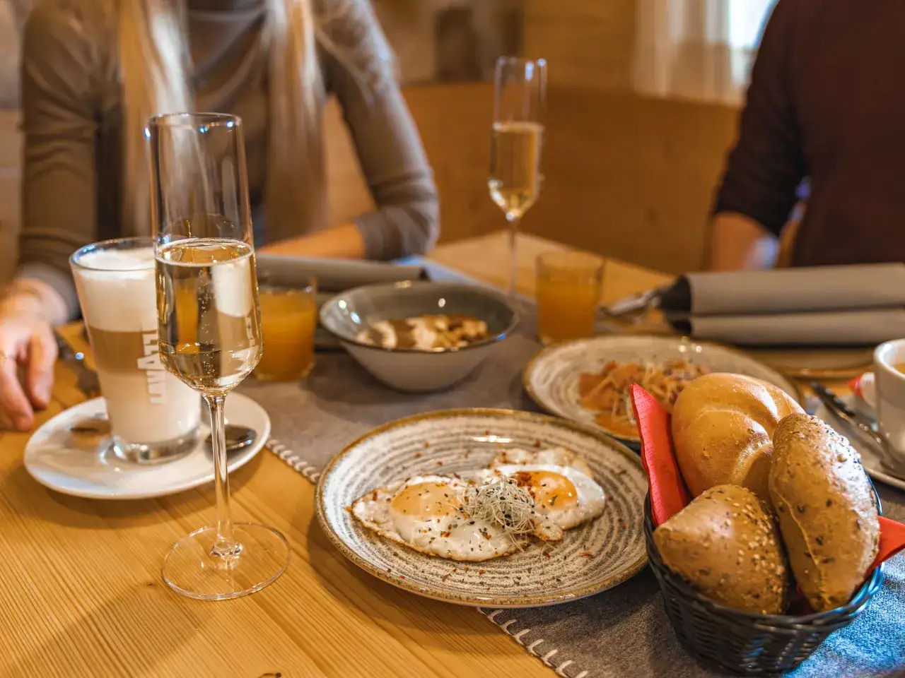 Frühstück mit Spiegeleiern, Brot und Sekt. Ein Glas Latte Macchiato und Orangensaft stehen bereit.