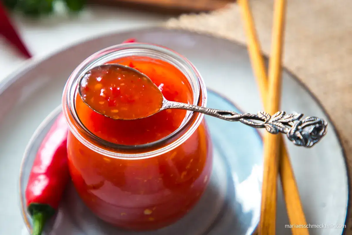 Selbstgemachte Chili-Sauce in einem Glas, mit einer L&ouml;ffelprobe und frischen Chilis. Perfekt f&uuml;r alle, die Chili-Sauce selber machen wollen.