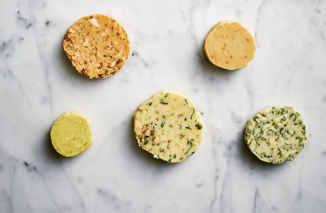 F&uuml;nf Scheiben Kr&auml;uterbutter, darunter eine mit Kr&auml;utern und Gew&uuml;rzen, die an Caf&eacute; de Paris Butter erinnert, liegen auf Marmor.