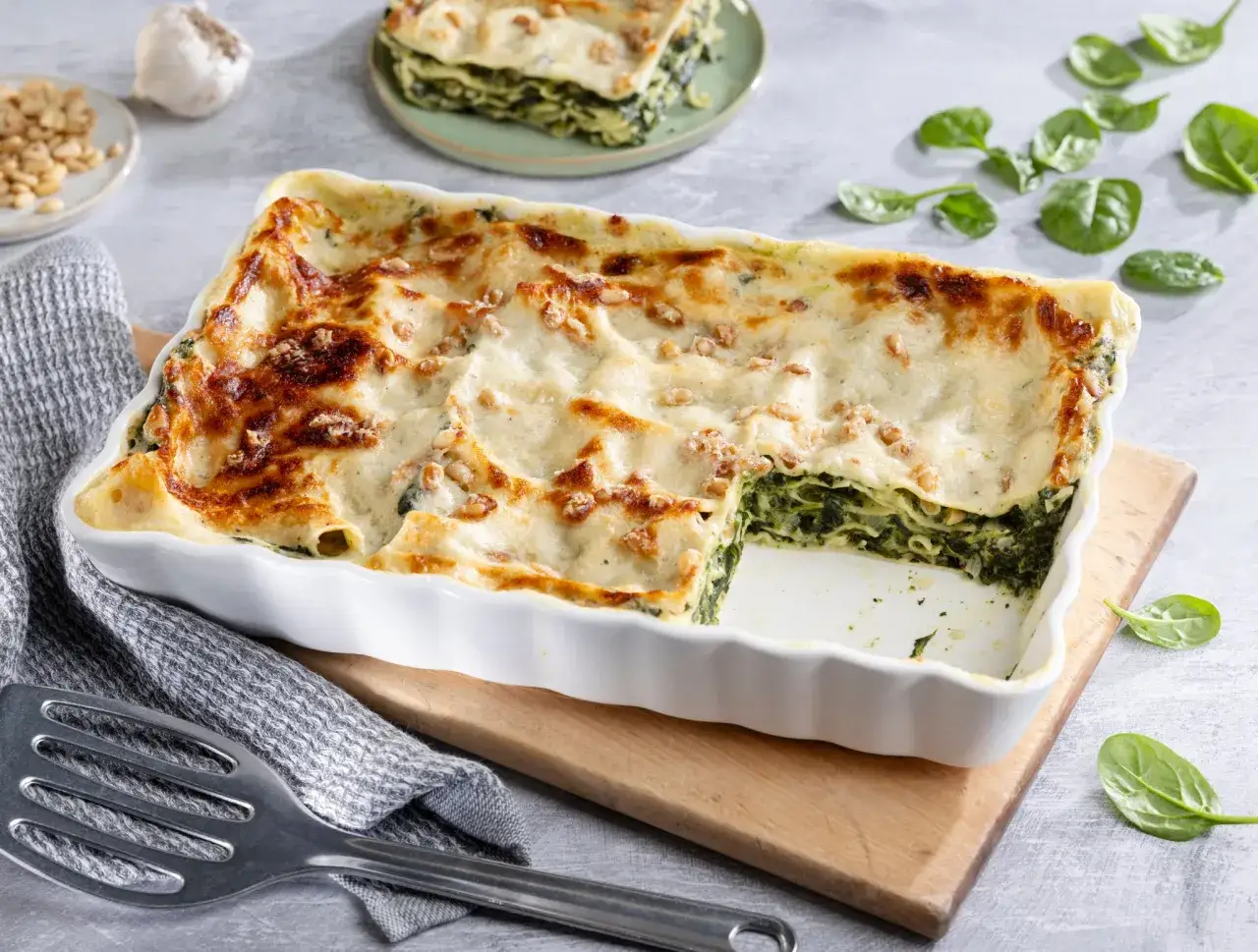 Herzhaftes Spinatlasagne-Rezept in einer Auflaufform, goldbraun überbacken. Frische Spinatblätter und Pinienkerne zieren die cremige Oberfläche.
