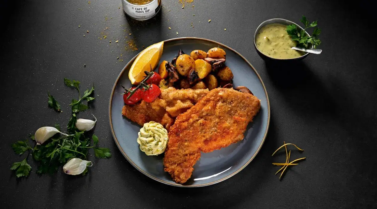 Ein knuspriger garnitur wiener schnitzel mit Bratkartoffeln, Tomaten, Zitrone und Kr&auml;uterbutter. Dazu eine Sauce und frische Kr&auml;uter.