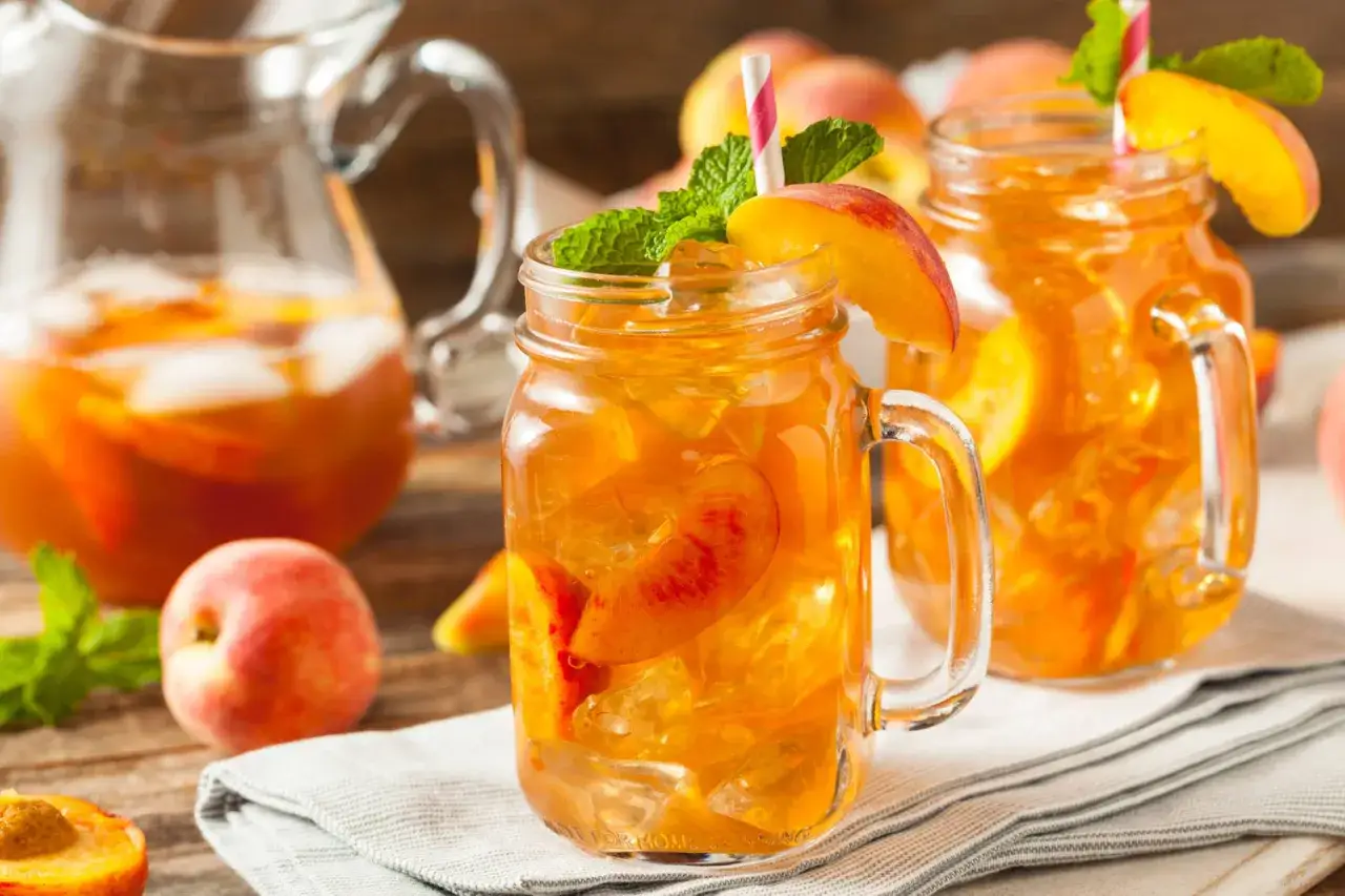 Erfrischender Pfirsich Eistee selber machen: Gl&auml;ser mit Eis, Pfirsichscheiben und Minze, daneben eine Karaffe mit dem leuchtend orangen Getr&auml;nk.