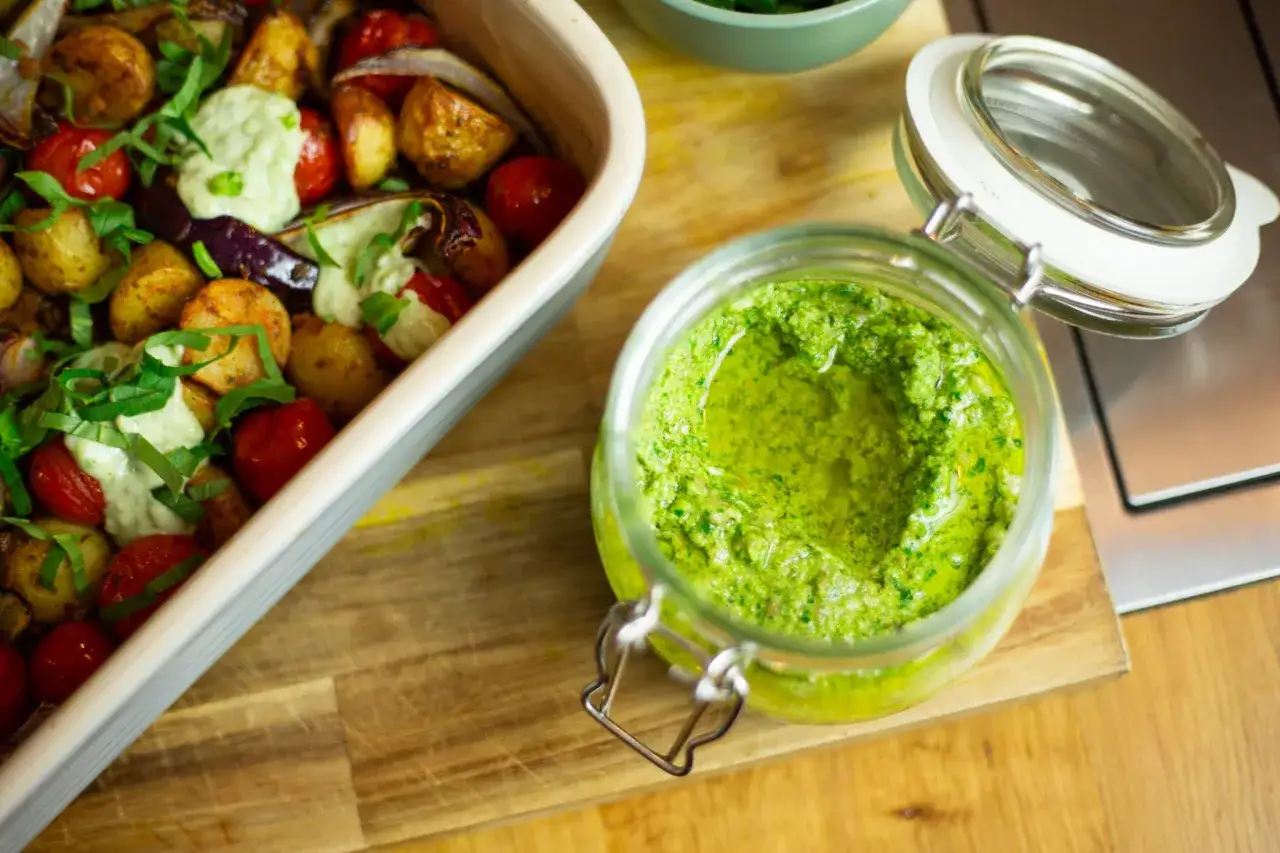 Frisches, gr&uuml;nes Pesto ohne Pinienkerne in einem Glas neben einem Auflauf mit Kartoffeln, Tomaten und Auberginen.