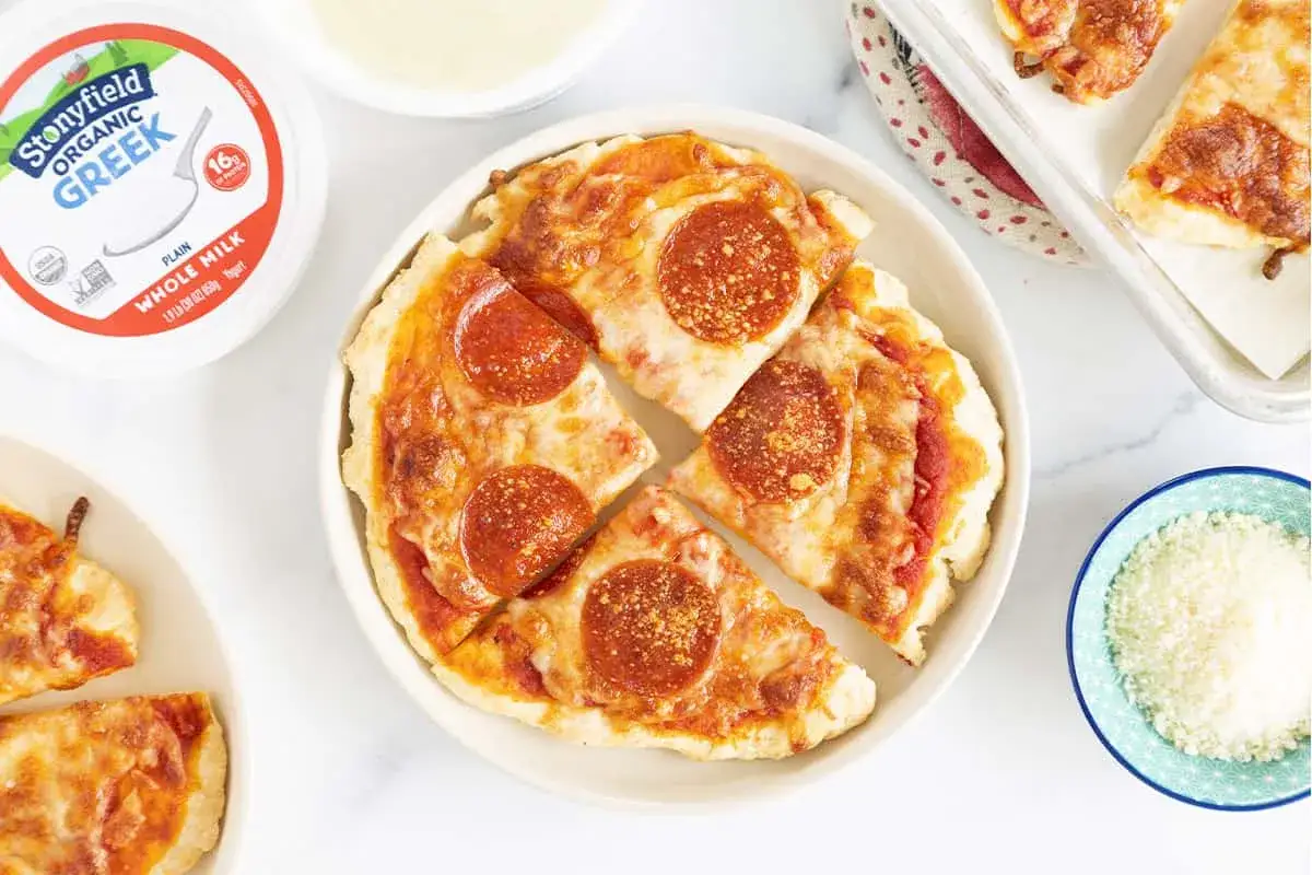 Pizzateig mit Joghurt als Basis für Mini-Pizzen mit Pepperoni. Daneben ein Becher griechischer Joghurt und geriebener Käse.