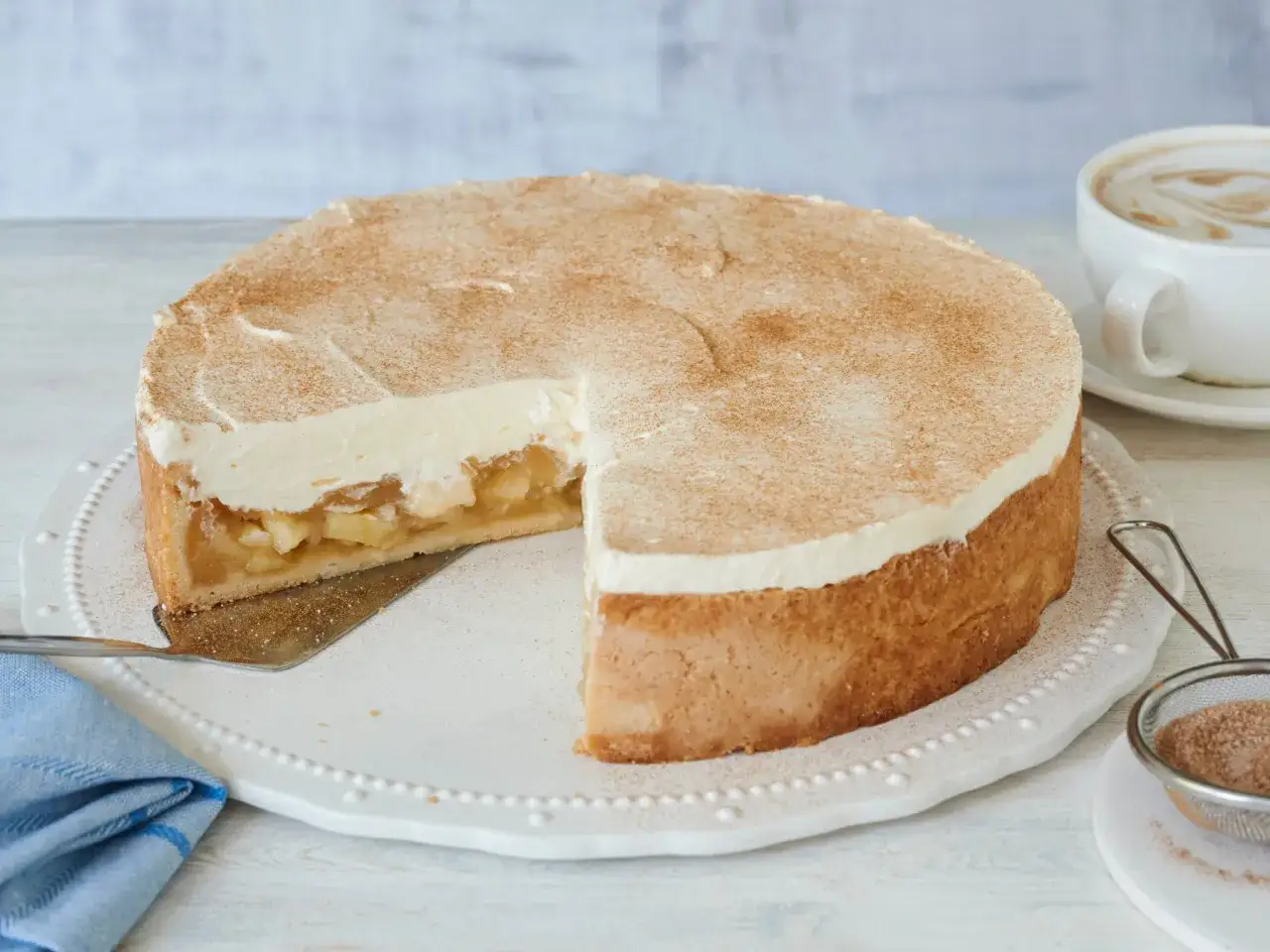 Saftige Apfel Schmand Torte mit Zimt best&auml;ubt, ein St&uuml;ck herausgeschnitten, daneben eine Tasse Cappuccino.