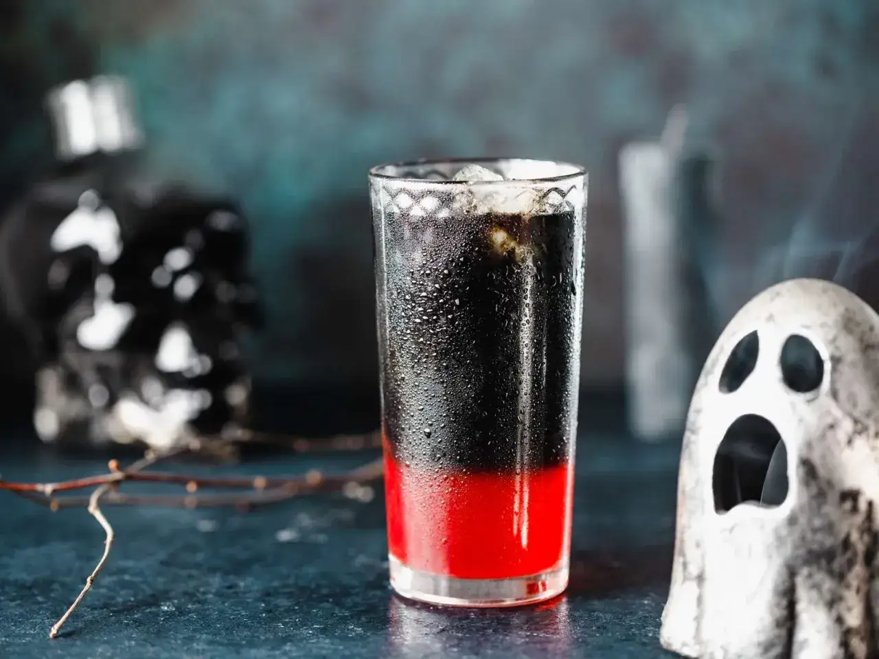 Gruseliger Drink mit roten und schwarzen Schichten, perfekt f&uuml;r Halloween Cocktails mit Alkohol. Ein Geist-Dekorst&uuml;ck und ein Totenkopf-Dekorst&uuml;ck im Hintergrund.
