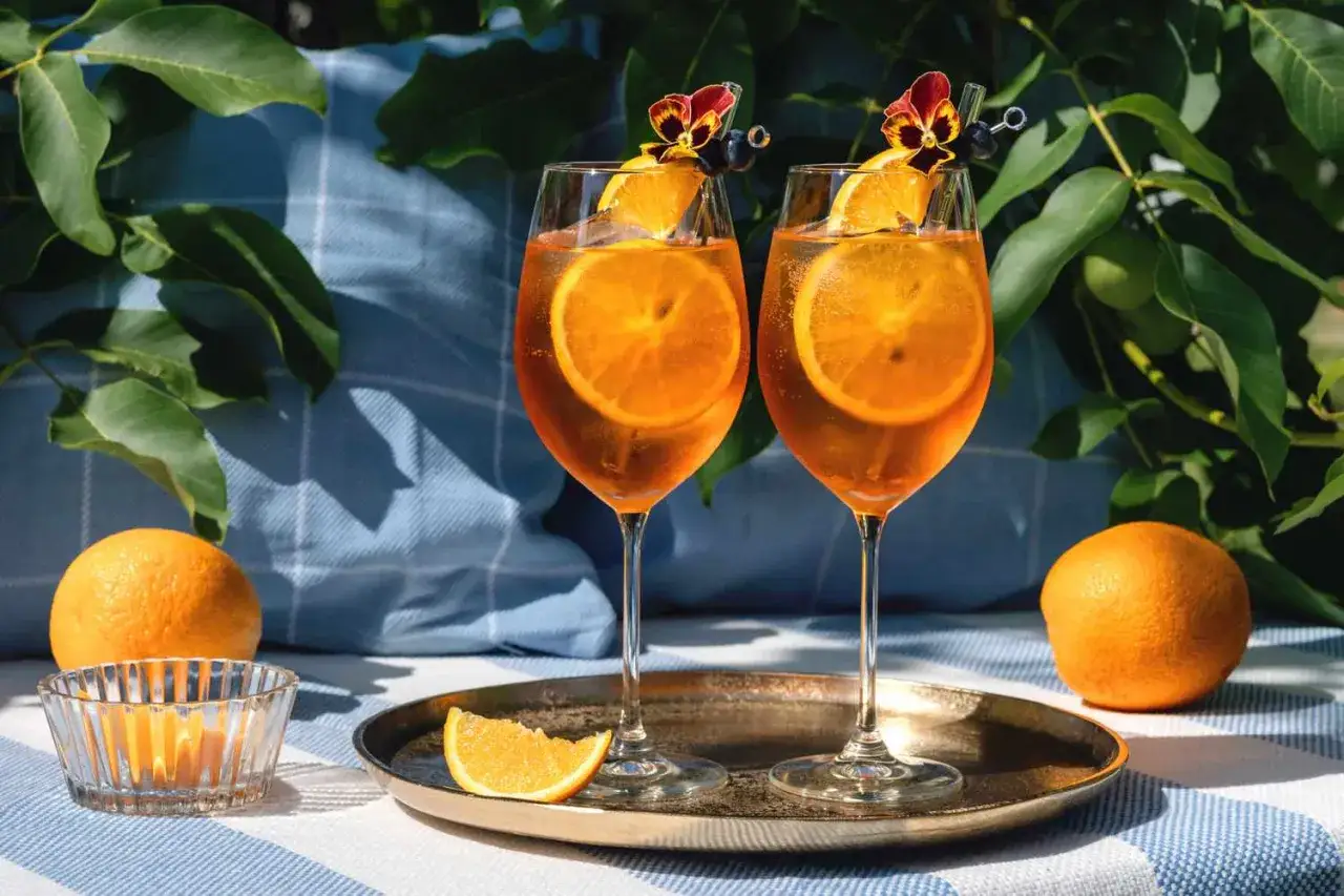 Zwei Aperol Spritz Gl&auml;ser mit Orangenscheiben und Blumen garniert, auf einem Tablett, umgeben von Orangen und gr&uuml;nen Bl&auml;ttern.