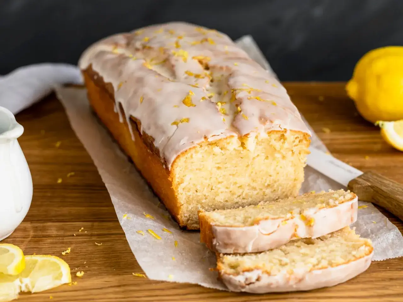 Saftiger veganer Zitronenkuchen mit Zuckerguss und Zitronenschale bestreut. Ein Stück ist angeschnitten, daneben liegen frische Zitronen.