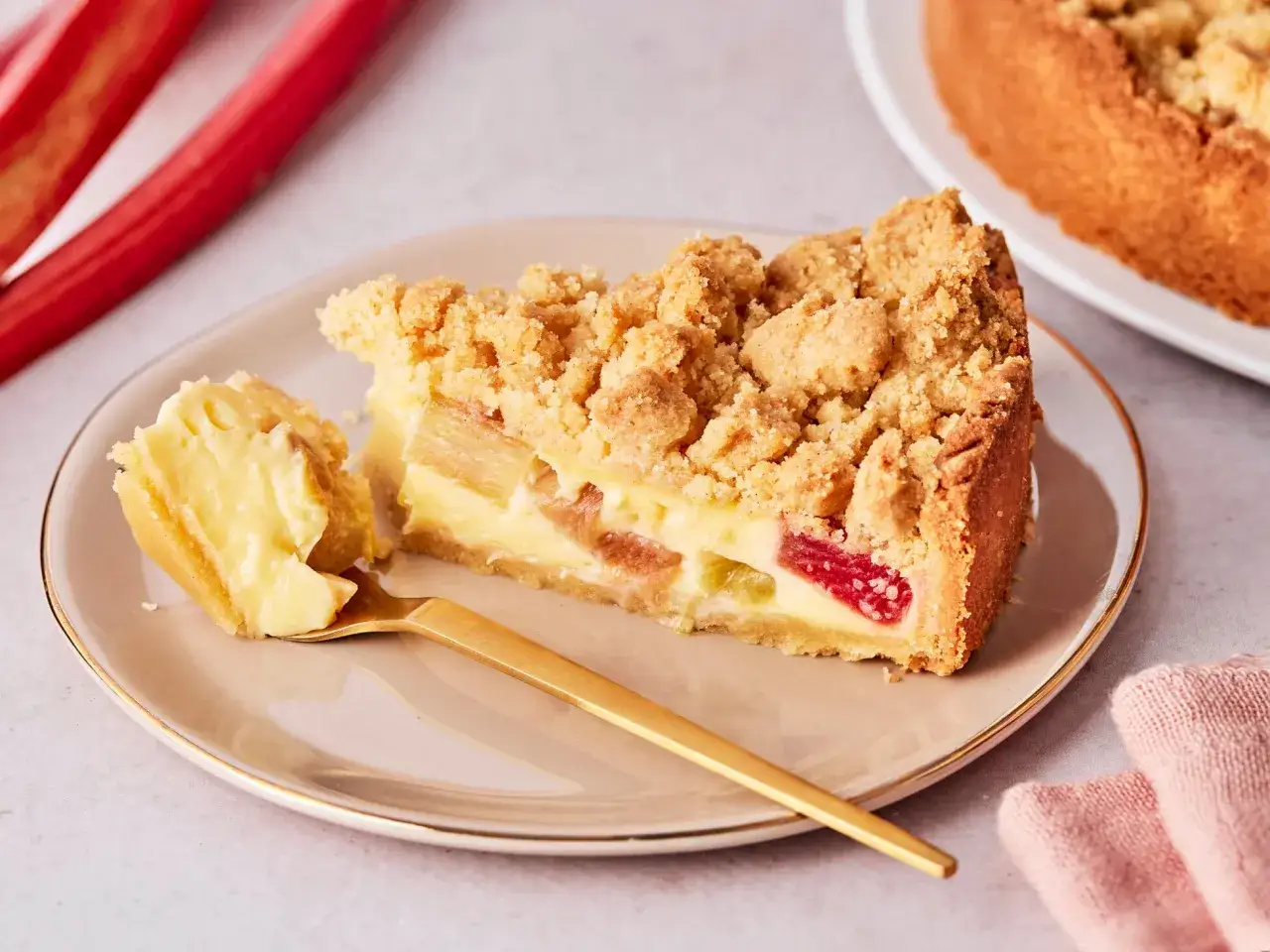 Ein St&uuml;ck Rhabarberkuchen mit Pudding und Streuseln auf einem Teller, daneben ein goldener L&ouml;ffel und frische Rhabarberstangen. Ein tolles Rhabarberkuchen Rezept mit Pudding!
