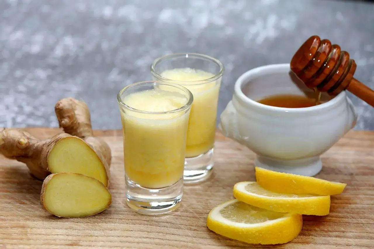 Zwei Gl&auml;ser mit gelbem Ingwer-Shot, daneben frischer Ingwer, Zitronenscheiben und Honig. Perfekt, um einen Ingwer Shot selber machen.
