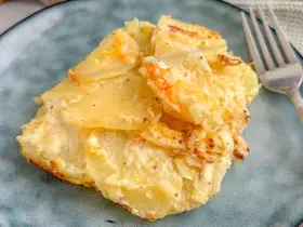 Ein Stück Kartoffelgratin einfach mit Sahne, goldbraun gebacken, auf einem blauen Teller. Daneben eine Gabel.