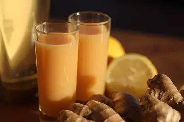 Zwei Gläser mit einem leuchtend orangen Ingwer Shot stehen neben frischem Ingwer und Zitronenscheiben.