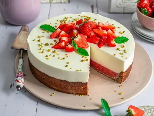 Erdbeer Joghurt Torte mit frischen Erdbeeren und Pistazien garniert. Ein Stück ist angeschnitten und zeigt die cremige Schicht.