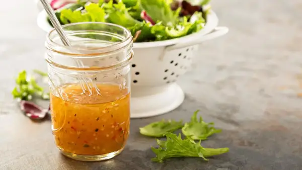 Ein Glas mit selbstgemachtem Salatdressing, das aussieht wie im Restaurant. Daneben ein Sieb mit frischem Salat.