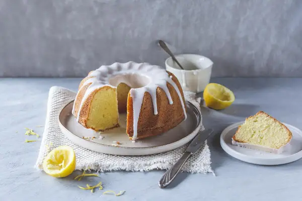 Ein saftiger Zitronenkuchen mit Zuckerguss, perfekt für ein Lemon Drizzle Cake Rezept. Daneben liegen frische Zitronen und ein Stück Kuchen.