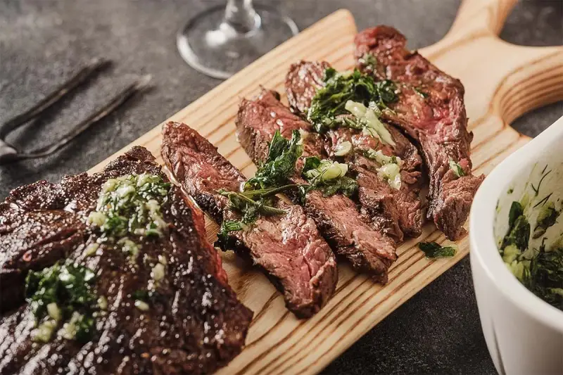 Saftiges, in Scheiben geschnittenes Flank Steak mit Kräutern und einer grünen Sauce auf einem Holzbrett serviert.