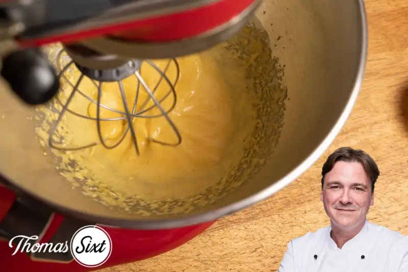 Küchenmaschine mixt eine cremige, gelbe Masse, die an Sauce Hollandaise erinnert. Ein Koch lächelt im Vordergrund.