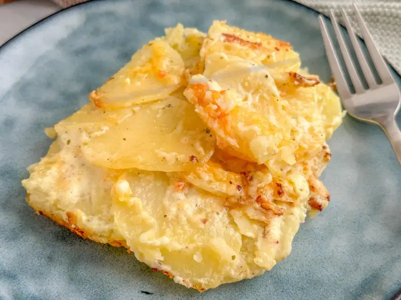 Ein Stück Kartoffelgratin einfach mit Sahne, goldbraun gebacken, auf einem blauen Teller. Daneben eine Gabel.