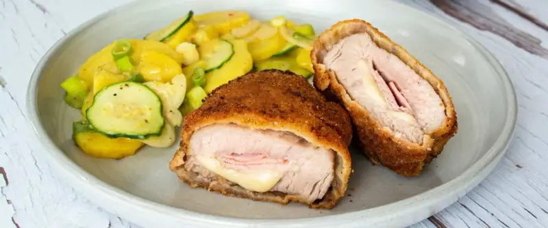 Zwei goldbraune Cordon-bleu-Schnitzel mit geschmolzenem Käse und Schinken, serviert mit einem Kartoffel-Zucchini-Salat.