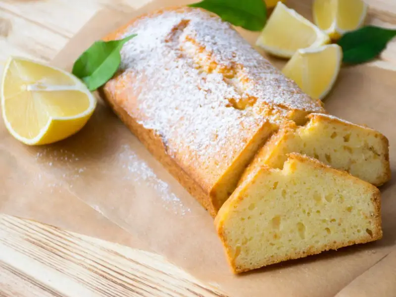 Saftiger veganer Zitronenkuchen, bestäubt mit Puderzucker, mit frischen Zitronenspalten und Blättern garniert.