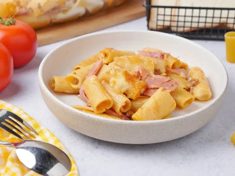 Herzhaftes Rigatoni al Forno mit Schinken und geschmolzenem Käse in einer Schüssel, daneben Tomaten und Brot.